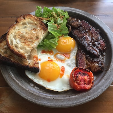 A hearty breakfast plate includes two sunny-side-up eggs with vibrant yellow yolks, crisp bacon strips, a slice of grilled tomato, fresh green lettuce, and two pieces of toasted sourdough bread. The plate is set on a wooden table, creating a rustic and inviting atmosphere.