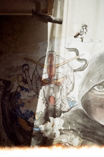 A painted mural on a wall depicting a robotic figure with a distinctive helmet and antenna-like structures. The wall shows signs of wear and decay, with parts of the paint peeling off. The mural is partially illuminated by a beam of sunlight coming from the top of the image. There is also a realistic eye painted on the wall next to the robotic figure, adding an intriguing contrast.