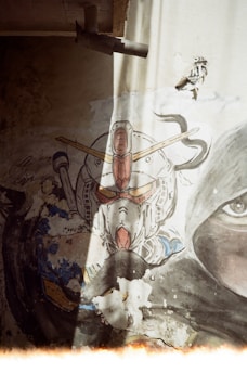 A painted mural on a wall depicting a robotic figure with a distinctive helmet and antenna-like structures. The wall shows signs of wear and decay, with parts of the paint peeling off. The mural is partially illuminated by a beam of sunlight coming from the top of the image. There is also a realistic eye painted on the wall next to the robotic figure, adding an intriguing contrast.