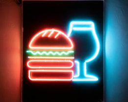 A brightly lit neon sign depicting a stylized hamburger with layers including lettuce and a drink next to it. The imagery is outlined in vibrant neon colors against a dark background.