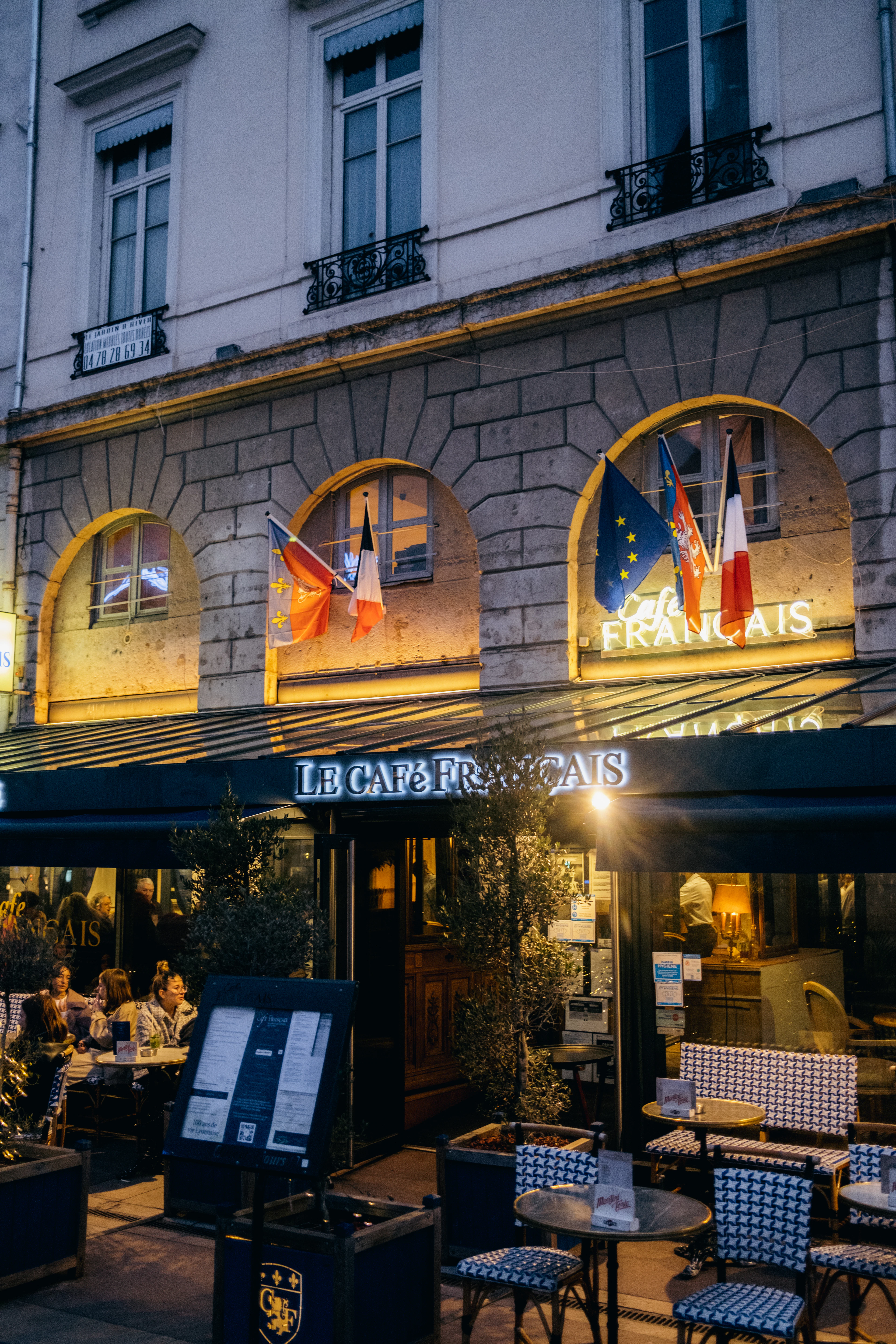 The glowing arched windows and lit sign of Le Cafe Francais brasserie in Lyon at dusk, diners at the pavement tables and empty bistro chairs set out under the awning.