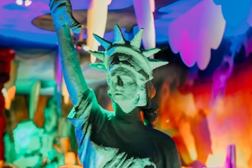 A stylized representation of the Statue of Liberty is set against a vibrant and colorful background, featuring a range of hues primarily in blue, pink, and orange. The statue is illuminated with green and turquoise lighting, giving it a surreal appearance.
