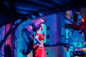 A stylized scene featuring a cartoonish, red-dressed figure posed in an animated manner. A dark, swirling form appears behind, while a vibrant red bird with exaggerated eyes and expression is in the foreground. The background consists of abstract, colorful architectural elements bathed in purple and blue lighting, giving a whimsical and playful atmosphere.