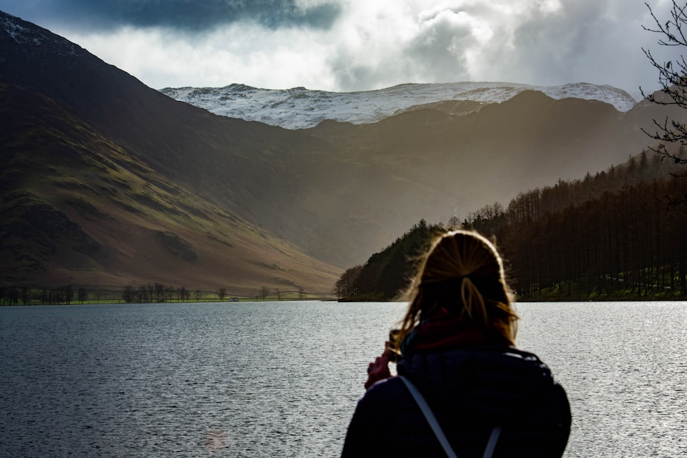 a woman standing in front of a lake with mountains in the background