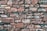 A wall made of irregularly shaped stones stacked closely together. The stones vary in size and have a rough, uneven texture with shades of brown and gray. Some stones have patches of lichen or moss, adding a natural element.