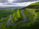 Wide-angle view of a winding road cutting through lush green hills under a dramatic sky.