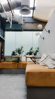 A cozy interior space featuring a seating arrangement with mustard-colored cushions. Above the seating area, there is a modern ceiling with exposed pipes and a duct system. The walls are adorned with simple white and black spherical wall lamps. The ambiance is enhanced by plants placed on the table and along the back wall, adding a touch of greenery.