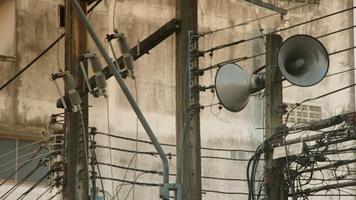 A complex network of electrical wires and equipment mounted on utility poles. Large loudspeakers are attached to the poles, along with various insulators and switches. The background features a weathered concrete wall, adding an industrial feel to the scene.