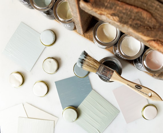 A collection of paint cans and sample swatches arranged on a white surface. There are several open jars of muted colors like pale pink, white, and light blue. A wooden-handled paintbrush with bristles that have traces of paint rests next to the cans. Some paint lids and sample cards lay scattered around, displaying the paint colors.
