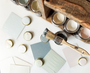 A collection of paint cans and sample swatches arranged on a white surface. There are several open jars of muted colors like pale pink, white, and light blue. A wooden-handled paintbrush with bristles that have traces of paint rests next to the cans. Some paint lids and sample cards lay scattered around, displaying the paint colors.