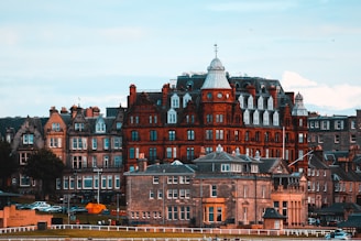 A row of historic buildings, featuring a prominent red-brick structure with a distinctive central dome and white window detailing, stand under a cloudy sky. In the foreground, a grassy area and some parked cars are visible, with a sign that reads 'World Golf Museum.'