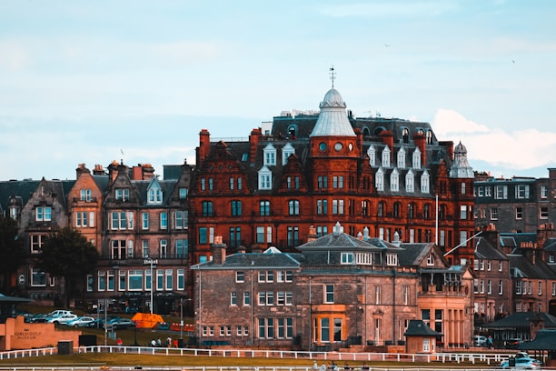 A row of historic buildings, featuring a prominent red-brick structure with a distinctive central dome and white window detailing, stand under a cloudy sky. In the foreground, a grassy area and some parked cars are visible, with a sign that reads 'World Golf Museum.'