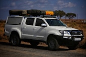 A white off-road vehicle with a robust build is parked on a road. The vehicle has a black front bull bar, side steps, and a roof rack holding camping gear, including a tent and a yellow item. The setting appears to be a dry, grassy landscape with a few scattered trees and partly cloudy skies.