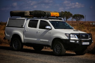 A white off-road vehicle with a robust build is parked on a road. The vehicle has a black front bull bar, side steps, and a roof rack holding camping gear, including a tent and a yellow item. The setting appears to be a dry, grassy landscape with a few scattered trees and partly cloudy skies.