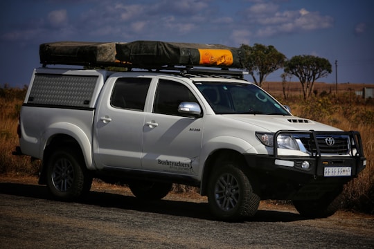 A white off-road vehicle with a robust build is parked on a road. The vehicle has a black front bull bar, side steps, and a roof rack holding camping gear, including a tent and a yellow item. The setting appears to be a dry, grassy landscape with a few scattered trees and partly cloudy skies.
