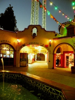 An outdoor plaza area illuminated with colorful lights, featuring a fountain surrounded by mosaic tiles in the foreground. The architecture displays a Spanish-style building with arched walkways and 'ARGADA' signage. Vendors and shops are visible through the arches, adding a festive atmosphere to the setting.