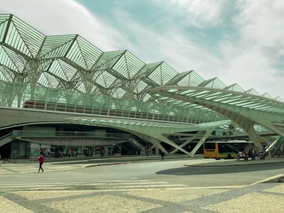 An architectural structure with a modern design features geometric patterns and extensive use of steel and glass. Passengers and vehicles like a bus are visible, emphasizing transportation. The sky in the background is partly cloudy.