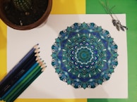 An intricately detailed mandala design colored in shades of blue and green is placed on a white sheet. Surrounding the mandala are several colored pencils that gradient from dark blue to green. To the left, there's a potted cactus, while on the right, some dried lavender is arranged. The background consists of a yellow and green backdrop.
