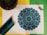 An intricately detailed mandala design colored in shades of blue and green is placed on a white sheet. Surrounding the mandala are several colored pencils that gradient from dark blue to green. To the left, there's a potted cactus, while on the right, some dried lavender is arranged. The background consists of a yellow and green backdrop.