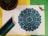 An intricately detailed mandala design colored in shades of blue and green is placed on a white sheet. Surrounding the mandala are several colored pencils that gradient from dark blue to green. To the left, there's a potted cactus, while on the right, some dried lavender is arranged. The background consists of a yellow and green backdrop.