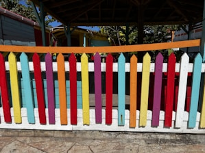 A colorful wooden picket fence painted in a variety of bright colors including red, yellow, blue, purple, and orange, standing in front of a shaded area under a pavilion structure. The background includes parts of a wooden building and foliage.