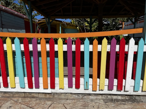 A colorful wooden picket fence painted in a variety of bright colors including red, yellow, blue, purple, and orange, standing in front of a shaded area under a pavilion structure. The background includes parts of a wooden building and foliage.