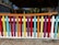 A colorful wooden picket fence painted in a variety of bright colors including red, yellow, blue, purple, and orange, standing in front of a shaded area under a pavilion structure. The background includes parts of a wooden building and foliage.