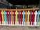 A colorful wooden picket fence painted in a variety of bright colors including red, yellow, blue, purple, and orange, standing in front of a shaded area under a pavilion structure. The background includes parts of a wooden building and foliage.