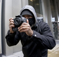 A person wearing a hoodie and face mask is holding a Canon camera, capturing a photograph. The setting appears to be urban with modern building structures.
