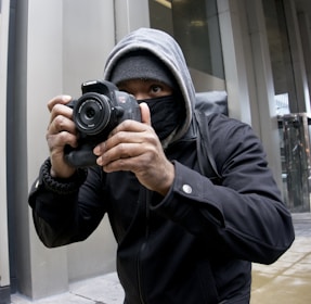 A person wearing a hoodie and face mask is holding a Canon camera, capturing a photograph. The setting appears to be urban with modern building structures.