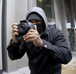 A person wearing a hoodie and face mask is holding a Canon camera, capturing a photograph. The setting appears to be urban with modern building structures.