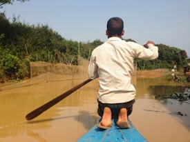 A person sits at the front of a boat holding a paddle, navigating through muddy waters surrounded by lush, green vegetation. The paddle is long and wooden, and the person's attire includes a light-colored shirt and dark pants. The scene conveys a sense of solitude and connection with nature.