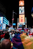 A large crowd of people gathers in a city square at night, surrounded by brightly lit billboards and digital screens. Many individuals are holding flags and signs, with a sense of unity and protest. The atmosphere is lively and bustling.
