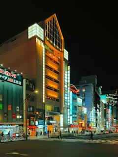 A bustling urban street scene at night with brightly lit buildings displaying a range of colorful advertisements and signs. The architecture suggests a vibrant city atmosphere with a mix of modern and slightly older structures. People can be seen walking on the sidewalks while vehicles travel on the roads, contributing to the lively ambiance.