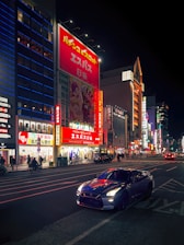 A nighttime urban scene featuring a brightly lit street lined with tall buildings adorned with colorful advertisements and neon lights. In the foreground, a sleek, modern sports car is parked on the road. Pedestrians and cyclists can be seen on the sidewalk, enjoying the vibrant city atmosphere.