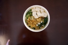 A white bowl is filled with a colorful array of ingredients including cubes of white tofu, finely chopped greens, and what appears to be fried shallots or onions.