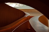 Curved, modern architecture with a polished wooden staircase gracefully winding through the interior. The soft lighting highlights the smooth textures and warm wood tones, creating an elegant and sophisticated atmosphere.