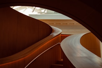 Curved, modern architecture with a polished wooden staircase gracefully winding through the interior. The soft lighting highlights the smooth textures and warm wood tones, creating an elegant and sophisticated atmosphere.