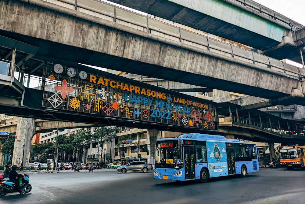 a blue bus driving down a street under a bridge