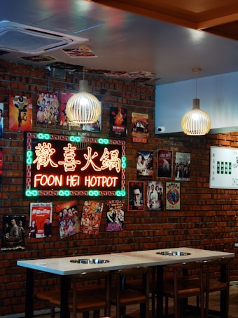 A neon sign with the text 'Foon Hei Hotpot' is mounted on a wall made of red bricks. The wall is decorated with various colorful posters, including scenes from movies and events. Two modern pendant lights hang from the ceiling above a table equipped with hot pot burners. The atmosphere is cozy and vibrant.