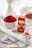 A white ceramic bowl is filled with vibrant red saffron threads placed on a rustic white wooden board. Nearby, a small red box labeled as premium saffron sits alongside a small white spoon containing some saffron threads. A cup of tea is placed in the background, next to a white ceramic teapot. Delicate purple and white flowers are scattered around the scene, adding a touch of elegance. The setting is bright and artfully arranged, suggesting a harmonious blend of culinary and aesthetic elements.