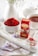 A white ceramic bowl is filled with vibrant red saffron threads placed on a rustic white wooden board. Nearby, a small red box labeled as premium saffron sits alongside a small white spoon containing some saffron threads. A cup of tea is placed in the background, next to a white ceramic teapot. Delicate purple and white flowers are scattered around the scene, adding a touch of elegance. The setting is bright and artfully arranged, suggesting a harmonious blend of culinary and aesthetic elements.