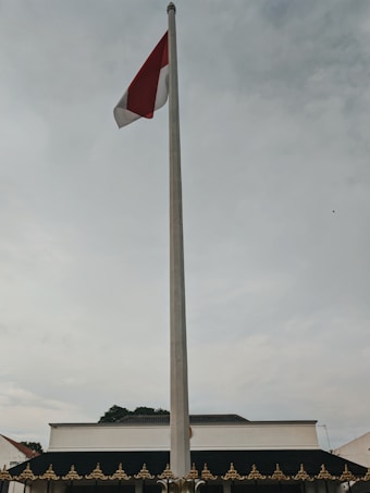 A tall flagpole stands prominently with a red and white flag waving in the breeze. The background features a traditional building with ornamental decorations along the roofline. The sky is overcast, with a mix of gray and soft white clouds.