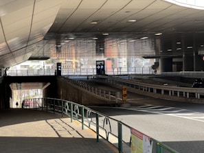 A modern urban underpass with tiled roof panels, two clear lanes for vehicles, and directional signs indicating parking. Pedestrian pathways are outlined with railings and a lone person in the distance. Light reflects off the surfaces, creating a dynamic interplay of shadow and color.