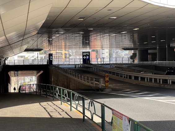 A modern urban underpass with tiled roof panels, two clear lanes for vehicles, and directional signs indicating parking. Pedestrian pathways are outlined with railings and a lone person in the distance. Light reflects off the surfaces, creating a dynamic interplay of shadow and color.