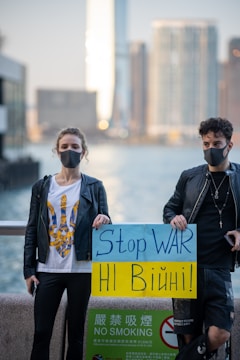 Two individuals wearing black face masks are standing outdoors in front of a blurred cityscape. One person holds a sign with the message 'Stop WAR' written in blue on a yellow and blue background. A 'NO SMOKING' sign with text in both English and Chinese is visible beneath.