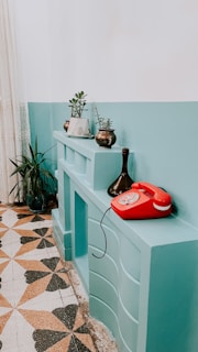 A vintage interior setting featuring a teal-painted shelf or mantle with potted plants on top and a red rotary telephone. The floor has a geometric patterned tile with earthy tones. A ceramic vase adds an artistic touch to the display.