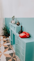 A vintage interior setting featuring a teal-painted shelf or mantle with potted plants on top and a red rotary telephone. The floor has a geometric patterned tile with earthy tones. A ceramic vase adds an artistic touch to the display.