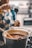 A person is holding a ladle with a thick, dark liquid pouring from it into a large pot filled with a rich, brown substance. The person is wearing an apron with a blue and yellow pattern. In the background, there is a slightly blurred kitchen setting.