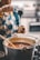 A person is holding a ladle with a thick, dark liquid pouring from it into a large pot filled with a rich, brown substance. The person is wearing an apron with a blue and yellow pattern. In the background, there is a slightly blurred kitchen setting.